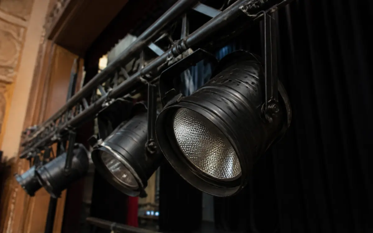 O serie de reflectoare negre de scenă, montate pe un sistem de grinzi metalice transversale (truss), într-o sală de spectacol cu o perdea neagră în fundal