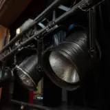 O serie de reflectoare negre de scenă, montate pe un sistem de grinzi metalice transversale (truss), într-o sală de spectacol cu o perdea neagră în fundal