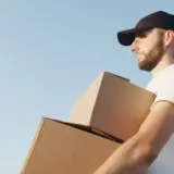 Un barbat cu barba, purtand o sapca neagra si un tricou alb, tine in brate doua cutii de carton pe fundalul unui cer albastru senin