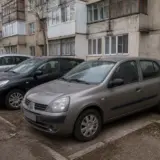 O mașină Renault Clio gri parcată în fața unui bloc de locuințe vechi, tip panel, cu mai multe alte autoturisme parcate în linie în fundal