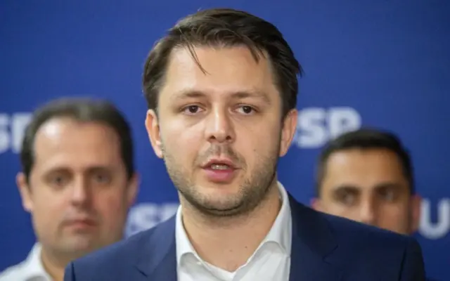 Un portret de tip plan mediu al viceprimarului Ruben Lațcău, vorbind în fața unui panou albastru