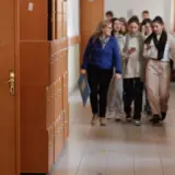 Un grup de elevi și un profesor merg pe un coridor de școală, pe lângă un rând de dulapuri metalice de culoare portocalie