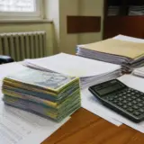 O stiva groasa de bancnote romanesti de 100 de lei (RON) asezata pe niste documente contabile, alaturi de un calculator negru pe un birou de lemn