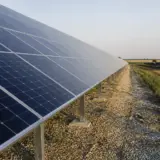 O serie de panouri solare montate pe rânduri pe un teren acoperit cu pietriș, într-o zonă rurală sau câmp deschis, sub un cer senin la apus sau răsărit