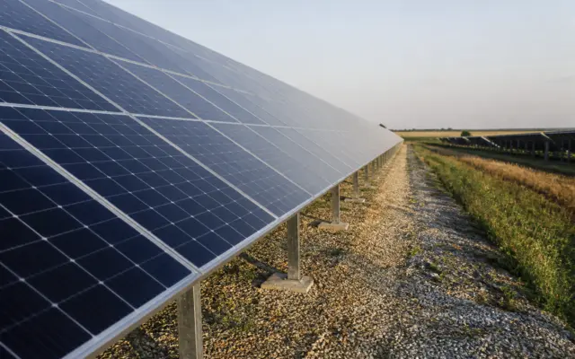 O serie de panouri solare montate pe rânduri pe un teren acoperit cu pietriș, într-o zonă rurală sau câmp deschis, sub un cer senin la apus sau răsărit