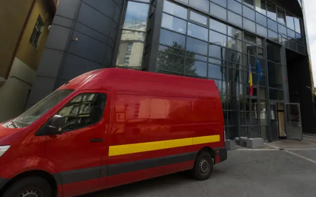 A red Poșta Română delivery van parked in front of a modern glass office building headquarters