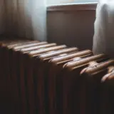 Un prim-plan al unui radiator (calorifer) vechi din fontă, situat sub o fereastră cu perdele albe transparente