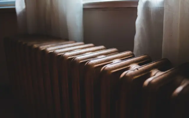 Un prim-plan al unui radiator (calorifer) vechi din fontă, situat sub o fereastră cu perdele albe transparente